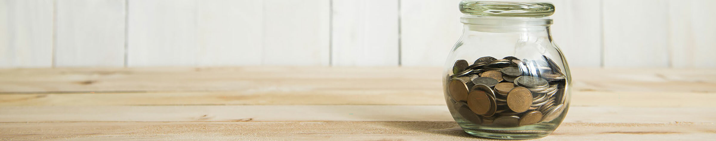 Small jar full of coins on a light wooden surface with a white wall