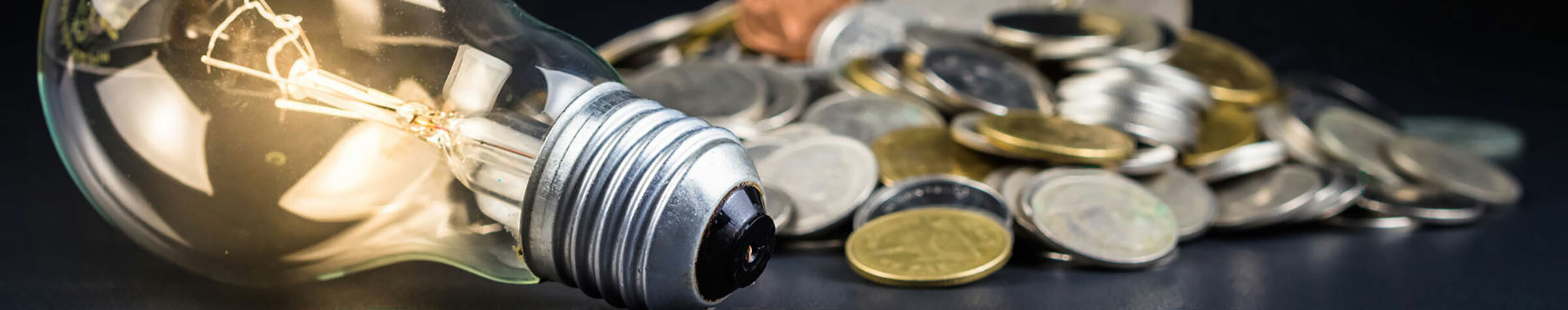 A lit lightbulb in front of a pile of coins on a dark surface